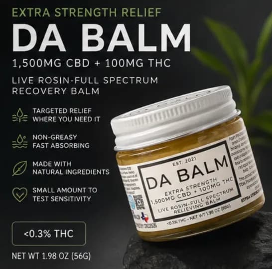The DA BALM jar on a dark charcoal background. A squat glass jar of olive-gold balm with a brushed silver screw lid sits on a piece of black stone, with a green plant frond blurred behind it. Large white type down the left gives the product name, the 1,500mg CBD and 100mg THC strength, and the 1.98 oz net weight.