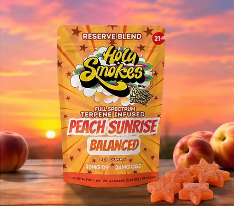 The orange Peach Sunrise Balanced pouch on a wooden table at sunrise, with whole peaches at the right and four sugared peach-colored star gummies in front.