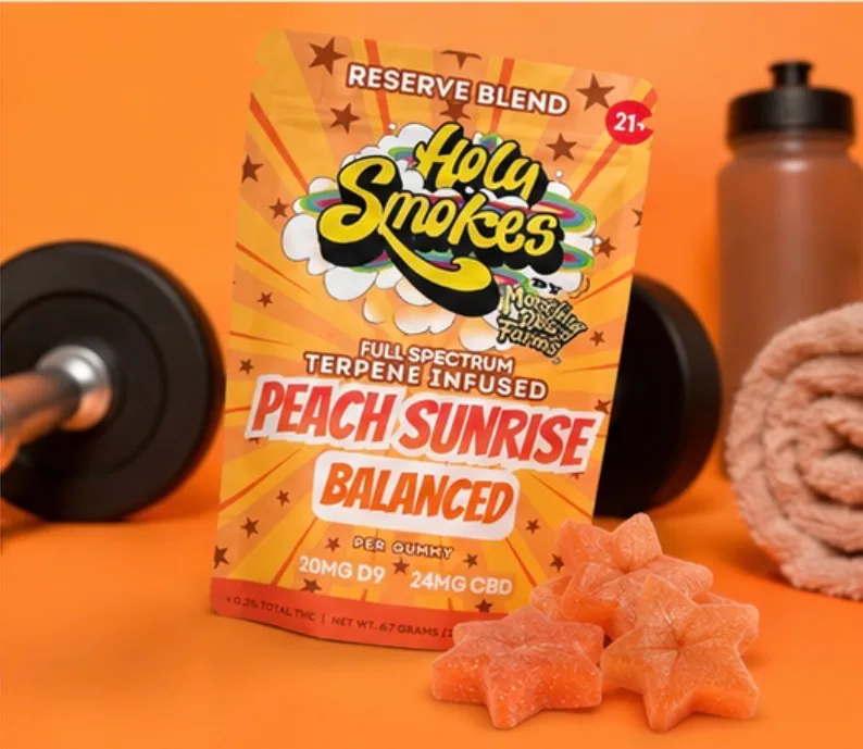 The pouch against a bright orange background with a dumbbell, a water bottle and a rolled towel behind it and four peach star gummies at the right.