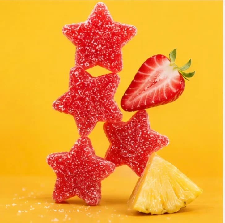 Five sugared red star gummies stacked in a loose tower against a yellow background, with half a strawberry and a wedge of pineapple leaning against them.